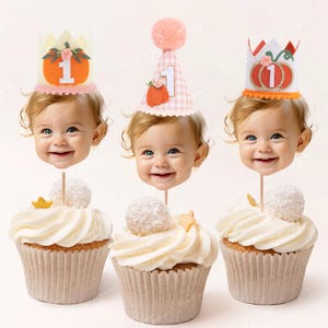 May include: Three cupcakes with white frosting and a single white ball on top. Each cupcake has a different topper: a pink and white gingham hat with a pumpkin and the number "1", a white crown with a pumpkin and the number "1", and a pink and white gingham hat with a pumpkin and the number "1".