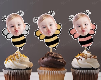 May include: Three cupcakes with bee-themed toppers featuring a baby's face. The bee designs have black and gold stripes, with silver, gold, and pink glitter wings. The cupcakes have white and chocolate frosting.