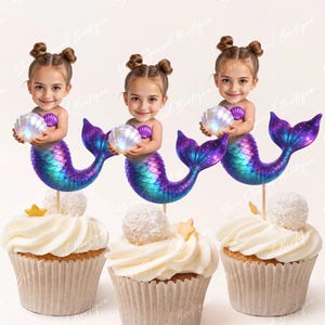 May include: Three cupcakes with mermaid decorations. Each cupcake has white frosting and a mermaid figure holding a pearl. The mermaid tails are blue and purple. The cupcakes are in paper cases. The image has the text "Your Special Boutique".