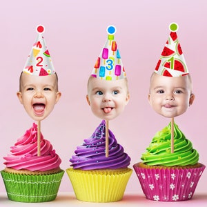 Op de afbeelding: Drie cupcakes met babygezichten op stokjes, elk met een feesthoedje. De cupcakes hebben roze, paarse en groene glazuur, met kleurrijke cupcakevormpjes. De hoeden hebben fruit- en ijslolly-ontwerpen, met de nummers 1, 2 en 3.
