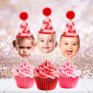 May include: Three cupcakes with pink frosting and red cupcake liners. Each cupcake has a photo of a child's face attached to a wooden stick. The children are wearing party hats with the number 2 and strawberry designs. A red pom-pom tops each hat.