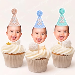 May include: Three cupcakes with white frosting, each topped with a baby face wearing a party hat with the number "2". The hats are in pastel colors: tan, blue, and teal, each with a matching pom-pom. A star and a white candy ball decorate each cupcake.