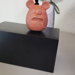 May include: A terracotta-colored ceramic ornament shaped like a mouse with a black cord hanging from the top. The ornament is sitting on a black box.