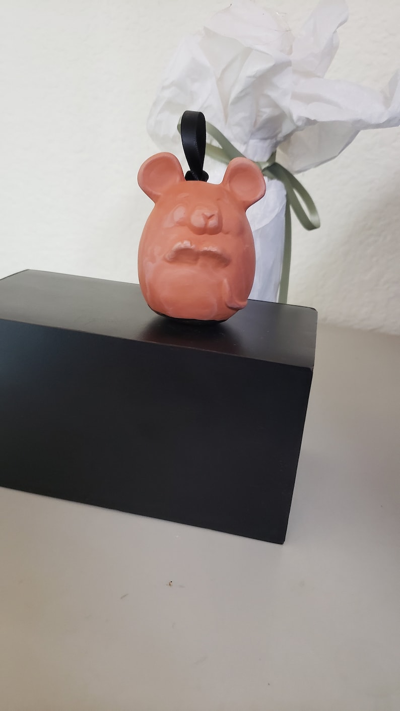 May include: A terracotta-colored ceramic ornament shaped like a mouse with a black cord hanging from the top. The ornament is sitting on a black box.