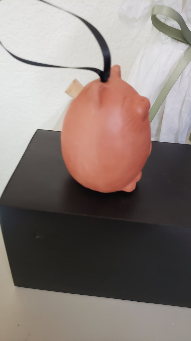 May include: A terracotta piggy bank ornament with a black ribbon and a wooden stopper. The piggy bank is sitting on a black box.