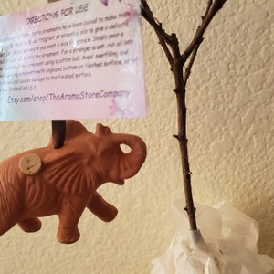 May include: Terracotta elephant ornament with a cork stopper on top. The ornament is hanging from a branch. The text on the tag reads "Directions for Use: These ornaments have been infused to make them... Etsy.com/shop/TheAromaStoreCompany"