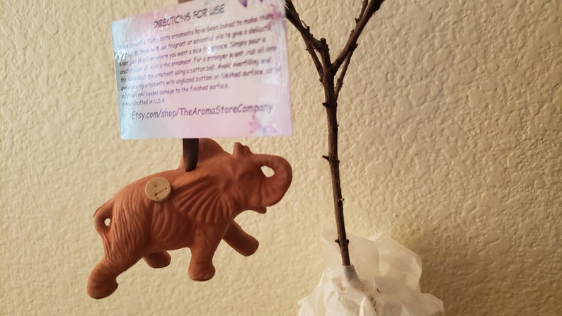 May include: Terracotta elephant ornament with a cork stopper on top. The ornament is hanging from a branch. The text on the tag reads "Directions for Use: These ornaments have been infused to make them... Etsy.com/shop/TheAromaStoreCompany"