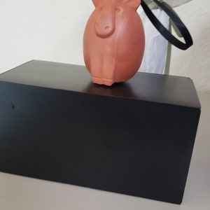 May include: A terracotta-colored ceramic ornament shaped like a capybara, hanging from a black ribbon. The ornament is sitting on top of a black box.