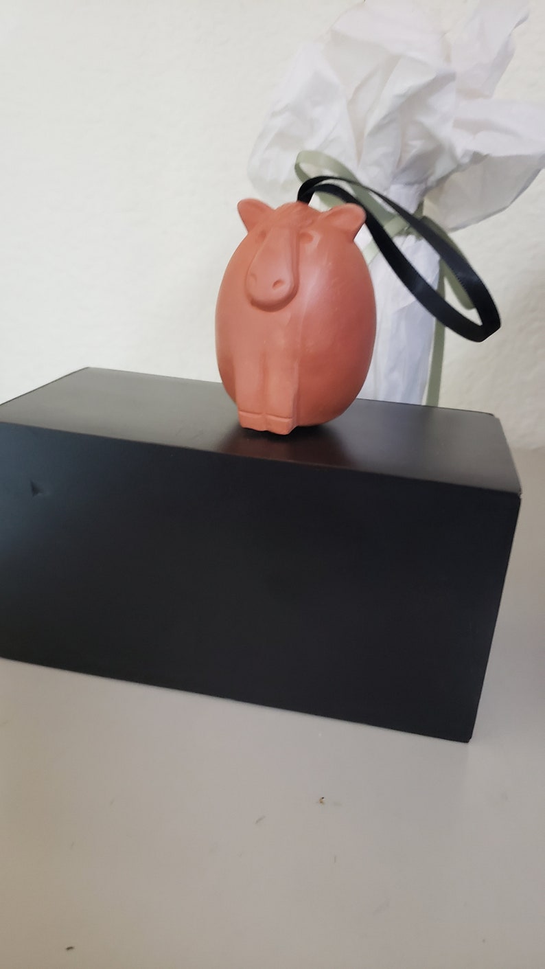 May include: A terracotta-colored ceramic ornament shaped like a capybara, hanging from a black ribbon. The ornament is sitting on top of a black box.