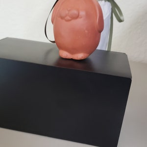 May include: A small, terracotta-colored ceramic ornament shaped like a dog with a black string attached. The ornament is sitting on a black box.