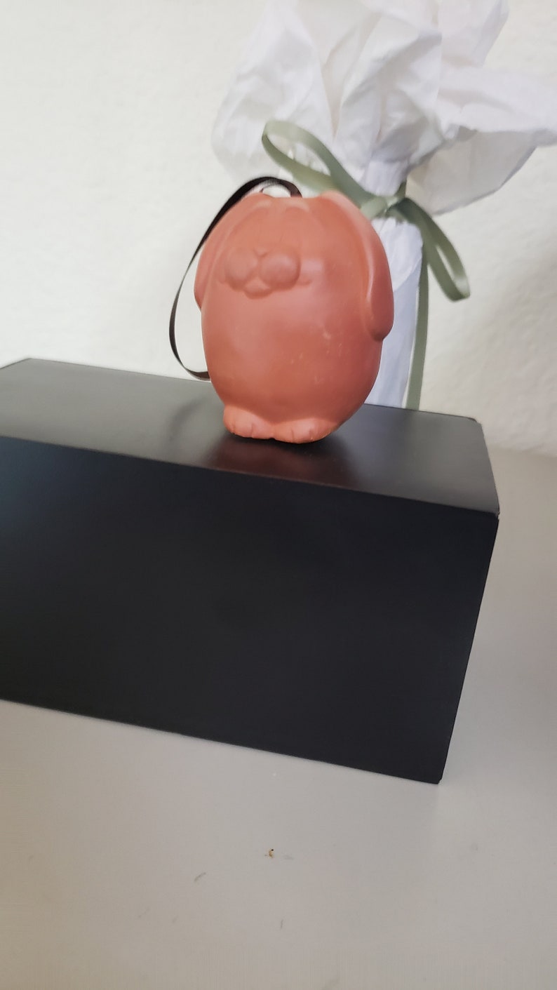 May include: A small, terracotta-colored ceramic ornament shaped like a dog with a black string attached. The ornament is sitting on a black box.