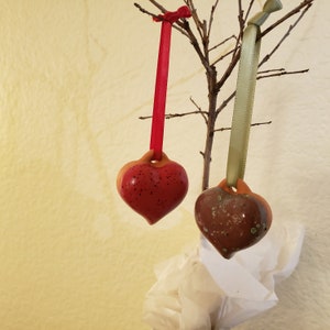 May include: Two ceramic heart ornaments hanging from a branch. The heart on the left is red with black speckles. The heart on the right is brown with green speckles.