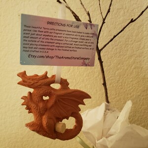 May include: A terracotta dragon ornament with a detailed design, hanging from a white ribbon. The ornament has a cork in the bottom and is displayed with a sprig of a tree and a card with instructions.
