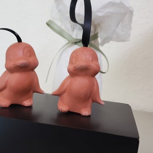 May include: Two terracotta bird ornaments with black ribbon hangers. The birds have a rounded body and a beak. They are standing on a black surface.