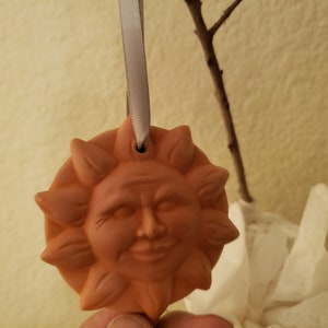 Puede incluir: Un adorno de sol de terracota con una cara sonriente y un agujero en la parte superior para colgar. El adorno est&aacute; sujeto a una cinta blanca.