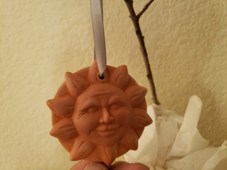 Puede incluir: Un adorno de sol de terracota con una cara sonriente y un agujero en la parte superior para colgar. El adorno est&aacute; sujeto a una cinta blanca.