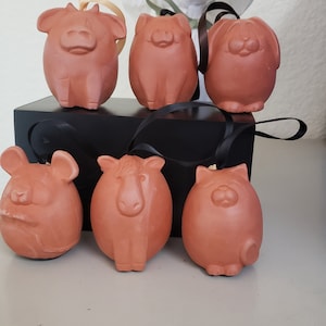 May include: Six terracotta ornaments shaped like different animals, including a cow, pig, rabbit, mouse, horse, and cat. The ornaments are hanging from a black ribbon and are displayed on a black surface.
