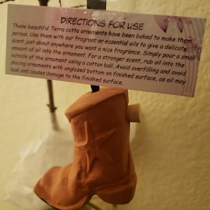 May include: A terracotta cowboy boot ornament with a star design. The ornament has a cork stopper and a tag with instructions for use. The tag reads "DIRECTIONS FOR USE".