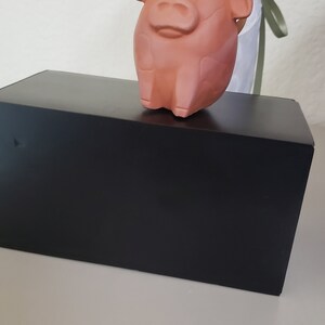 May include: A terracotta-colored ceramic cow figurine with a white ribbon around its neck. The cow is standing on a black box.
