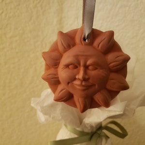 Puede incluir: Un adorno de sol de terracota con una cara sonriente y un agujero para colgar. El adorno est&aacute; atado con una cinta gris.