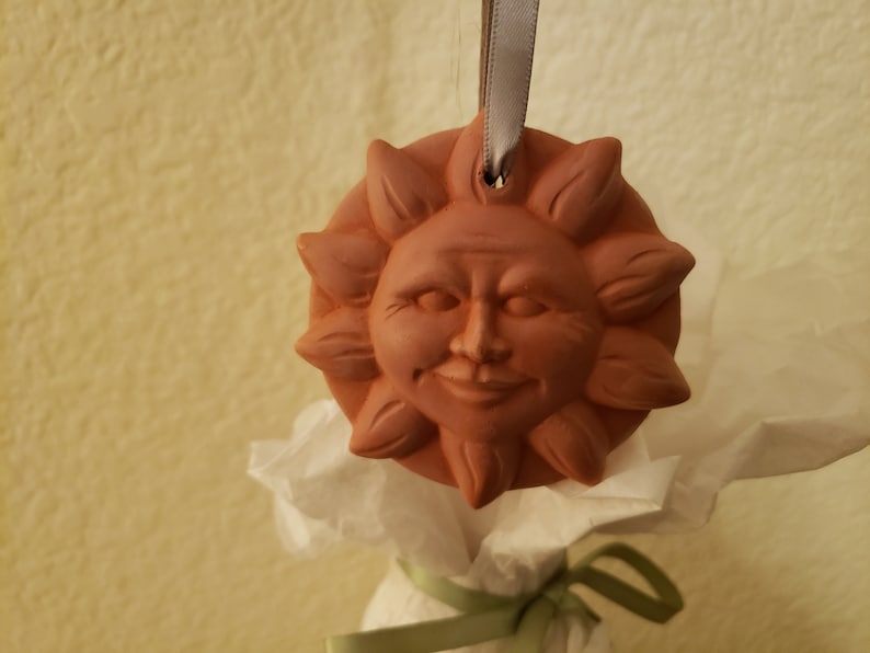 Puede incluir: Un adorno de sol de terracota con una cara sonriente y un agujero para colgar. El adorno est&aacute; atado con una cinta gris.