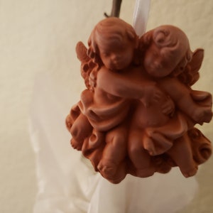May include: Two terracotta angels with wings, hugging each other. The angels are painted in a light brown color and have a rustic look.