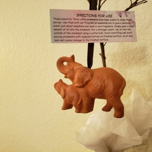 May include: A terracotta ornament of two elephants, one larger than the other, with a brown ribbon attached. The ornament is hanging from a branch. The text on the card reads: "DIRECTIONS FOR USE These beautiful Terra cotta ornaments have been baked to make them porous. Use them with our fragrant or essential oils to give a delicate scent just about anywhere you want a nice fragrance. Simply pour a small amount of oil into the ornament. For a stronger scent, rub oil into the outside of the ornament using a cotton ball. Avoid overfilling and avoid placing ornaments with unglazed bottom on finished surface, as oil may leak and causes damage to the finished surface."