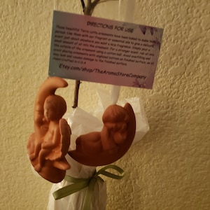 May include: Two terracotta angel figurines, one sitting and one lying down, are attached to a crescent moon. The figurines are a light brown color and have a rustic finish. The moon is also terracotta and has a rough texture. The figurines are hanging from a branch with a white ribbon and a green ribbon tied around the branch. A small card with the text "DIRECTIONS FOR USE" is attached to the branch. The card also includes the text "Etsy.com/shop/TheAromaStoreCompany".