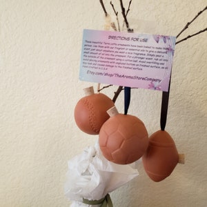 May include: Three terracotta ornaments shaped like a baseball, soccer ball, and football, each with a cork stopper and a dark ribbon for hanging. A card with instructions is visible. The ornaments are hand-crafted.