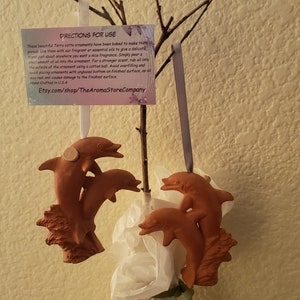 May include: Two terracotta dolphin ornaments hanging from a branch. The dolphins are in a jumping pose. A small card with instructions for use is attached to the branch. The card reads "Instructions for Use: These beautiful Terra cotta ornaments have been hand-baked to make them unique. Just heat with your finger or a disc of essential oil, gently rub the oil about the ornament with an unglazed surface. Simply pour a small amount of the essential oil on a cotton ball and rub it on the outside of the ornament. For a stronger scent, swirling and gently placing the ornament upside down, avoid over-heating as it may crack and cause damage to the finished surface. Hand-Crafted in U.S.A. Etsy.com/shop/TheAromaStoreCompany"