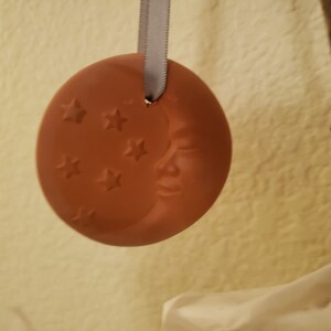 Puede incluir: Un adorno de cer&aacute;mica de color terracota con forma de media luna con una cara y estrellas. El adorno est&aacute; colgado de una cinta blanca.