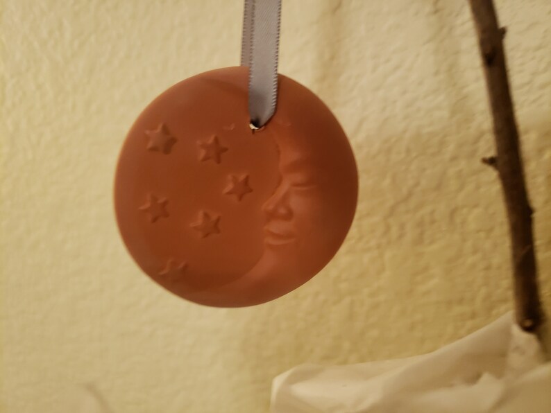 Puede incluir: Un adorno de cer&aacute;mica de color terracota con forma de media luna con una cara y estrellas. El adorno est&aacute; colgado de una cinta blanca.