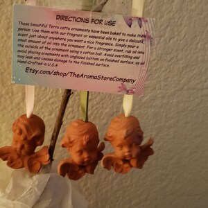 May include: Three terracotta angel ornaments with wings, hanging from a branch. The ornaments are a light brown color and have a rustic, handmade look. A small card with instructions for use is attached to the ornaments. The card reads "Directions for Use" and includes instructions for using essential oils with the ornaments. The card also includes the website Etsy.com/shop/TheAromaStoreCompany.