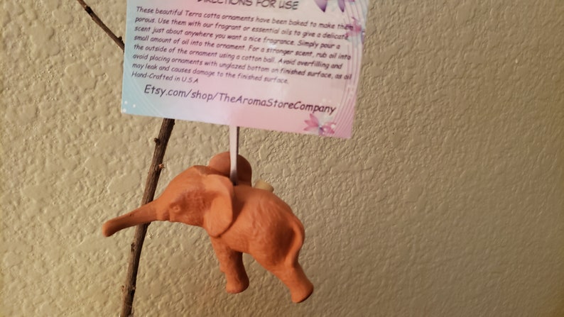 May include: A terracotta elephant ornament with a hole in the top for adding essential oils. The ornament is hanging from a branch. The text on the card reads: "These beautiful Terra cotta ornaments have been baked to make them porous. Use them with our fragrant or essential oils to give a delicate scent just about anywhere you want a nice fragrance. Simply pour a small amount of oil into the ornament. For a stronger scent, rub a little on the outside of the ornament using a cotton ball. Avoid overfilling and avoid placing ornaments with unglazed bottom on finished surface, as oil may leak and causes damage to the finished surface. Hand-Crafted in USA Etsy.com/shop/TheAromaStoreCompany"