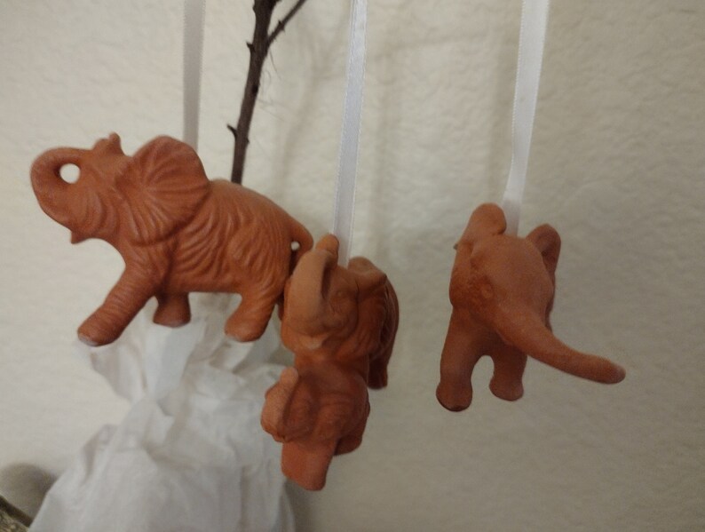 May include: Three terracotta elephant ornaments hanging from white strings. The elephants are all facing different directions and have a rustic, hand-crafted look.