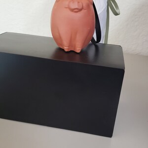 May include: A terracotta pig figurine with a black string attached to its head. The pig is sitting on a black box.