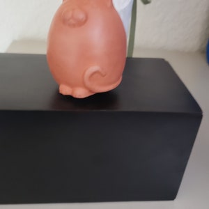 May include: A terracotta-colored ceramic ornament shaped like a cat. The ornament is hanging from a black ribbon and is sitting on a black box.