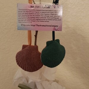 May include: Two terracotta shell-shaped ornaments, one is a deep green and the other is a light brown. The ornaments are hanging from a branch. A sign with directions for use is attached to the branch. The sign reads: "DIRECTIONS FOR USE These beautiful Terra cotta ornaments have been baked to make them porous. Use them with our fragrant or essential oils to give a delicate scent just about anywhere you want a nice fragrance. Simply pour a small amount of oil into the ornament. For a stronger scent, rub oil into the outside of the ornament using a cotton ball. Avoid overfilling and avoid placing ornaments with unglazed bottom on finished surface, as oil may leak and causes damage to the finished surface. Hand-Crafted in USA Etsy.com/shop/TheAromaStoreCompany"