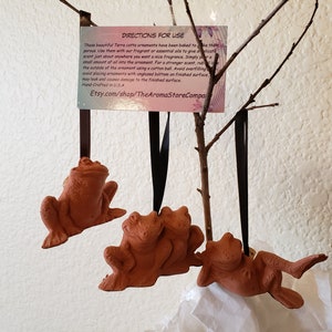 May include: Three terracotta frog ornaments hanging from black ribbon. The frogs are in various poses, including sitting and laying down. The ornaments are hand-crafted in the USA. The image includes directions for use: "These beautiful Terra cotta ornaments have been baked to...