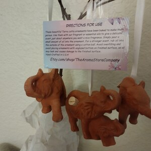 May include: Four terracotta elephant ornaments with cork stoppers. The ornaments are hanging from a white ribbon. The ornaments are brown and have a rustic finish. The text on the card reads: "DIRECTIONS FOR USE These beautiful Terra cotta ornaments have been baked to make them porous. Use them with our fragrant or essential oils to give a delicate scent just about anywhere you want a nice fragrance. Simply pour a small amount of oil into the ornament. For a stronger scent, rub oil into the outside of the ornament using a cotton ball. Avoid overfilling and avoid placing ornaments with unglazed bottom on finished surface, as oil may leak and causes damage to the finished surface. Hand-Crafted in USA Etsy.com/shop/TheAromaStoreCompany"