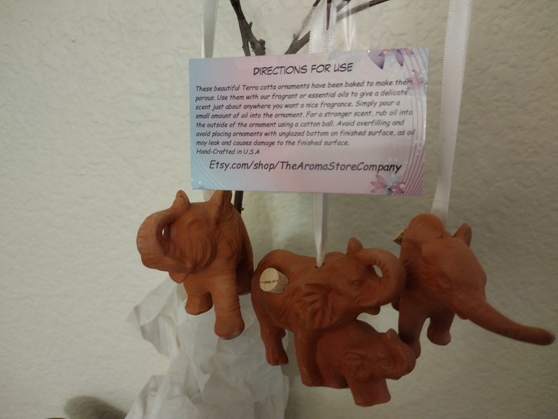 May include: Four terracotta elephant ornaments with cork stoppers. The ornaments are hanging from a white ribbon. The ornaments are brown and have a rustic finish. The text on the card reads: "DIRECTIONS FOR USE These beautiful Terra cotta ornaments have been baked to make them porous. Use them with our fragrant or essential oils to give a delicate scent just about anywhere you want a nice fragrance. Simply pour a small amount of oil into the ornament. For a stronger scent, rub oil into the outside of the ornament using a cotton ball. Avoid overfilling and avoid placing ornaments with unglazed bottom on finished surface, as oil may leak and causes damage to the finished surface. Hand-Crafted in USA Etsy.com/shop/TheAromaStoreCompany"