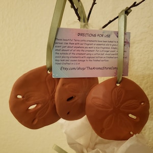 May include: Three terracotta sand dollar ornaments with a tag that reads "Directions for Use" and includes instructions for using essential oils. The tag also includes the website Etsy.com/shop/TheAromaStoreCom.