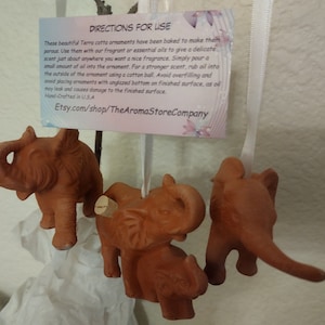 May include: Four terracotta elephant ornaments with cork stoppers. The ornaments are hanging from white ribbon. A card with instructions for use is attached to the ribbon. The card reads "Directions for Use" and includes instructions for using essential oils with the ornaments. The card also includes the Etsy shop name "Etsy.com/shop/TheAromaStoreCompany".