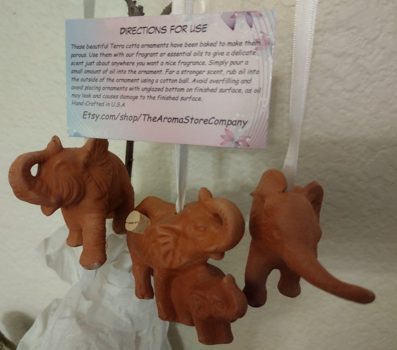 May include: Four terracotta elephant ornaments with cork stoppers. The ornaments are hanging from white ribbon. A card with instructions for use is attached to the ribbon. The card reads "Directions for Use" and includes instructions for using essential oils with the ornaments. The card also includes the Etsy shop name "Etsy.com/shop/TheAromaStoreCompany".
