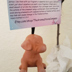 May include: A terracotta dog ornament, suspended by a black cord, is the focal point. A card with instructions is visible. The ornament is hand-crafted in the USA and designed to hold essential oils for fragrance.