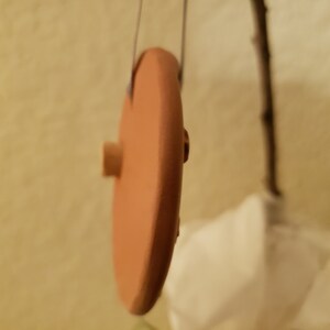 Puede incluir: Un disco redondo de color terracota con un peque&ntilde;o pomo de madera en el lateral. El disco est&aacute; colgado de una cuerda.