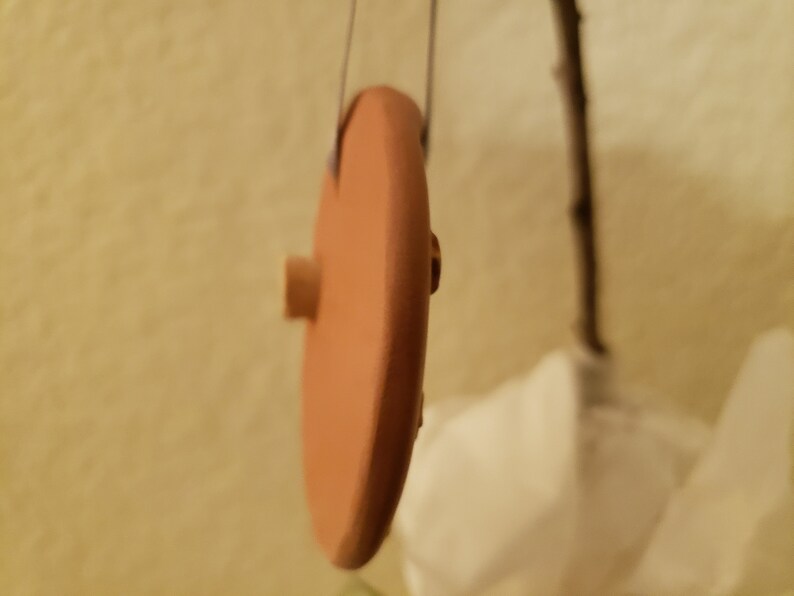 Puede incluir: Un disco redondo de color terracota con un peque&ntilde;o pomo de madera en el lateral. El disco est&aacute; colgado de una cuerda.