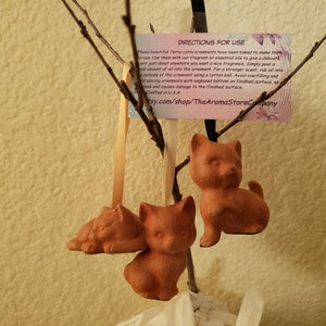 May include: Three terracotta cat ornaments hanging from a branch. The ornaments are a light brown color and have a rustic, handmade look. The ornaments are perfect for adding a touch of whimsy to any home.