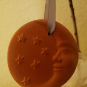 Puede incluir: Un adorno de cer&aacute;mica de color terracota con forma de luna creciente con una cara sonriente y cinco estrellas. El adorno est&aacute; colgado de una cinta blanca.