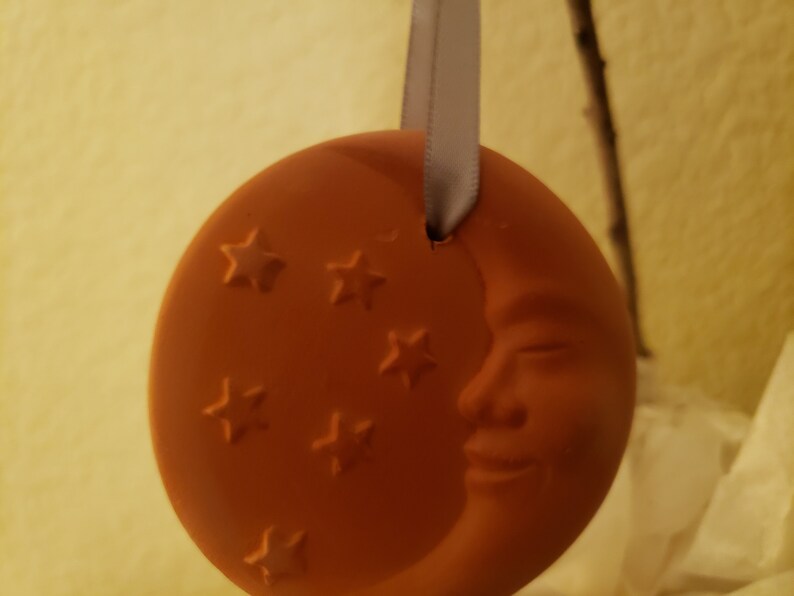 Puede incluir: Un adorno de cer&aacute;mica de color terracota con forma de luna creciente con una cara sonriente y cinco estrellas. El adorno est&aacute; colgado de una cinta blanca.